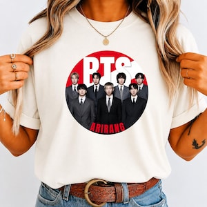 May include: A cream-colored t-shirt featuring a circular graphic with a red background. Inside the circle, seven individuals in dark suits are arranged in a group. The text BTS is printed in large white letters above them, and ARIRANG is in red.