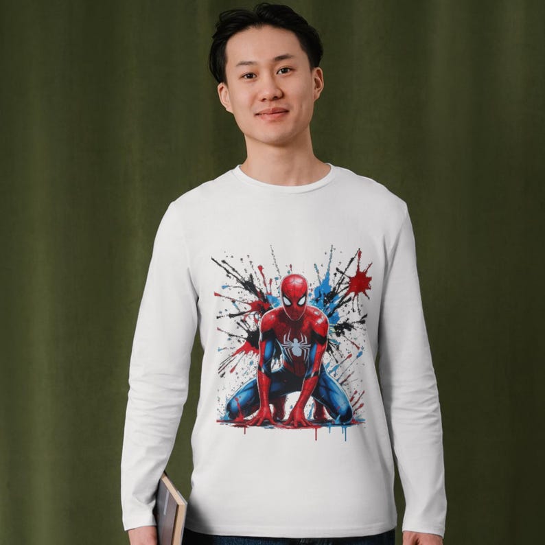 May include: A white long-sleeve shirt featuring a graphic print of a red and blue superhero character in a crouching pose. The design includes vibrant paint splatter effects in red, blue, and black surrounding the central figure on the chest.