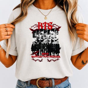 May include: A light gray t-shirt featuring a graphic print with the text BTS and BANGTAN SONYEONDAN. The design includes black and white portraits of seven individuals arranged in vertical panels, surrounded by red barbed wire and bird motifs.