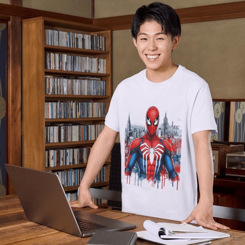 May include: A white short-sleeved t-shirt featuring a graphic print of the superhero Spider-Man in his red and blue suit with a white spider emblem on the chest, set against a stylized cityscape background with red and blue paint drip accents.