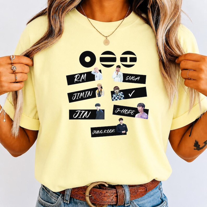 May include: A pale yellow t-shirt featuring a graphic print with three black circular icons at the top. Below are seven individual photographs of people arranged in rows, each paired with the names RM, SUGA, JIMIN, JIN, J-HOPE, and JUNG KOOK.