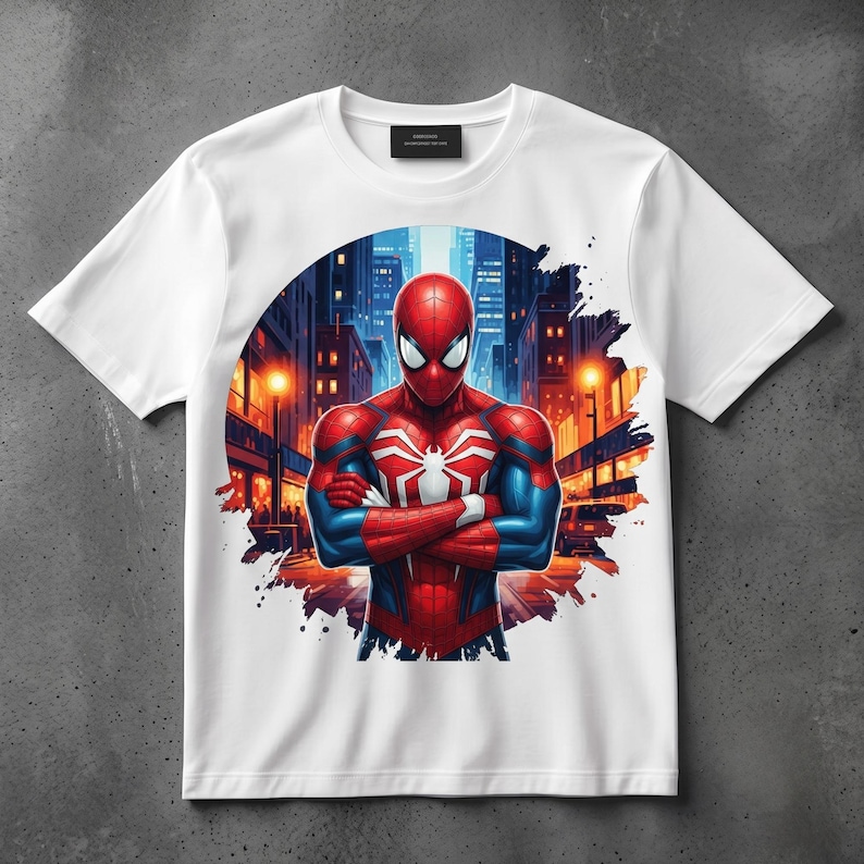 May include: A white short-sleeve t-shirt featuring a circular graphic print of a red and blue superhero with a white spider emblem on the chest. The character stands with arms crossed against a backdrop of a city street at night with glowing lights.