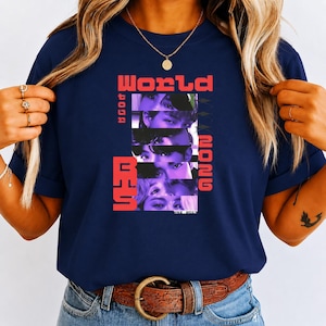 May include: A navy blue short-sleeve t-shirt featuring a graphic print with the text "WORLD" and "2026" in red, alongside a vertical stack of five cropped facial images in purple tones and the text "BTS" in red, with "EST. 2013" at the bottom.