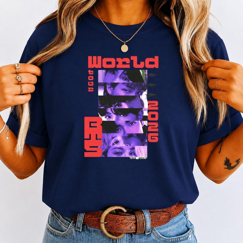 May include: A navy blue short-sleeve t-shirt featuring a graphic print with the text "WORLD" and "2026" in red, alongside a vertical stack of five cropped facial images in purple tones and the text "BTS" in red, with "EST. 2013" at the bottom.