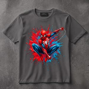 May include: A charcoal gray short-sleeve t-shirt featuring a central graphic of a red and blue costumed superhero in a crouching pose. The character is surrounded by a vibrant red and blue paint splatter effect with a thin white web line.