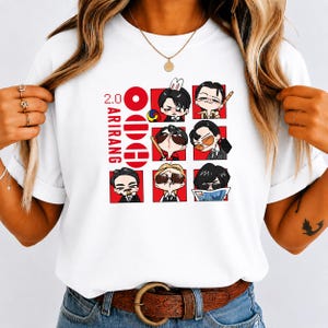 May include: A white t-shirt featuring a graphic print with the text 2.0 ARIRANG. The design includes a vertical column of three red circles and a grid of six cartoon character illustrations in various poses, wearing sunglasses and formal attire.