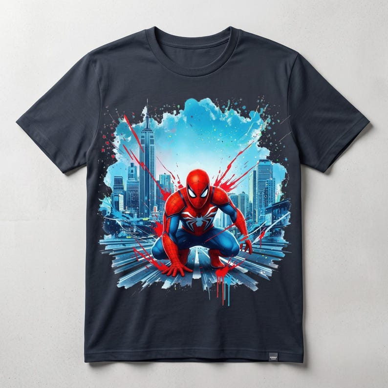 May include: A dark gray short-sleeve t-shirt featuring a graphic print of a red and blue costumed character in a crouching pose. The background displays a stylized city skyline with blue and red paint splatter effects across the chest area.