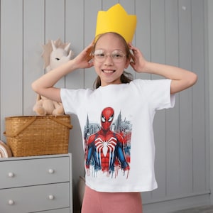 May include: A white short-sleeved t-shirt featuring a graphic print of the superhero Spider-Man in a red and blue suit with a white spider emblem on the chest, set against a detailed cityscape background with red and blue paint splatter accents.