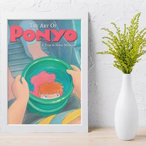 May include: A framed art print titled The Art of Ponyo, A Film by Hayao Miyazaki. The illustration depicts a character holding a green bucket containing a small orange-haired figure. The print is displayed in a white frame next to a white vase.