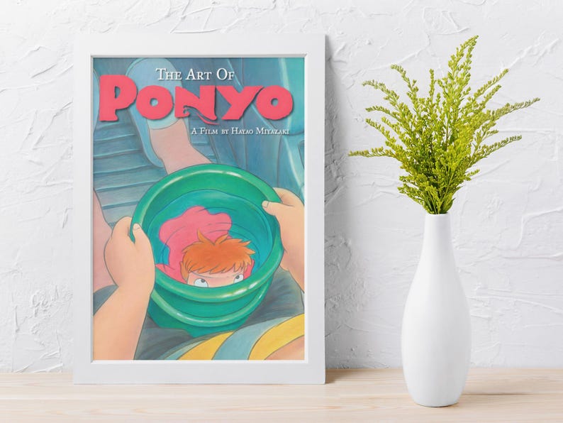 May include: A framed art print titled The Art of Ponyo, A Film by Hayao Miyazaki. The illustration depicts a character holding a green bucket containing a small orange-haired figure. The print is displayed in a white frame next to a white vase.