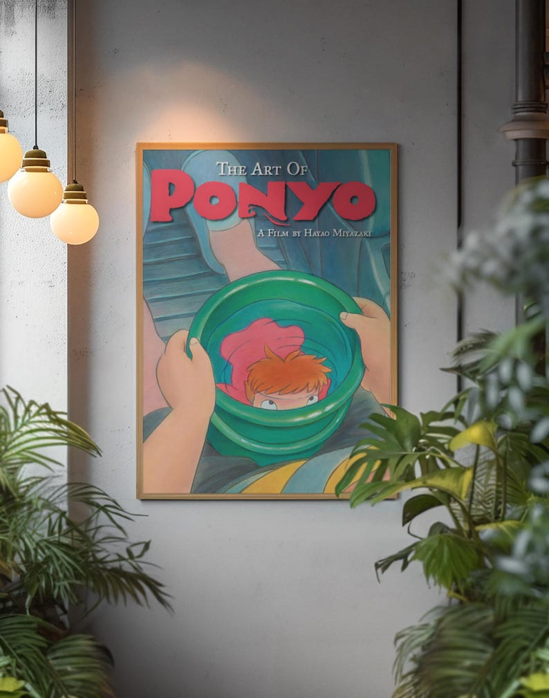 May include: A rectangular framed poster titled The Art Of Ponyo A Film By Hayao Miyazaki. The illustration depicts a person holding a green bucket containing a small orange-haired character. The text is printed in bold red and white lettering.