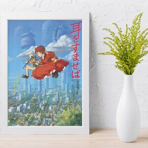 May include: A framed poster featuring anime artwork. The image depicts two characters in mid-air over a cityscape, with text in Japanese. The artwork is set against a blue sky with cloud-like structures. A white vase with green foliage is to the right.