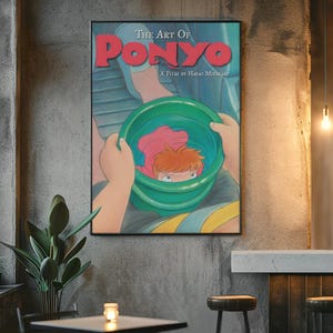 May include: A framed poster titled The Art of Ponyo, A Film by Hayao Miyazaki. The illustration depicts a character with orange hair inside a green bucket, held by hands against a blue background. The text is printed in bold red and white.