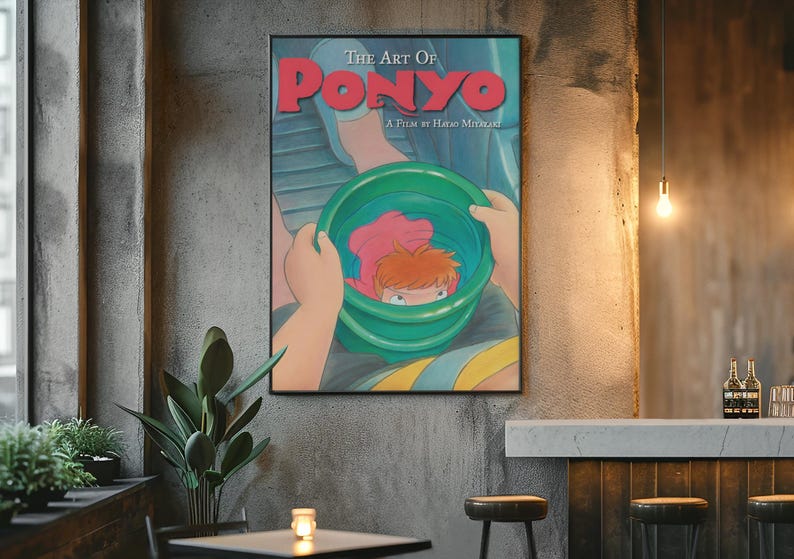 May include: A framed poster titled The Art of Ponyo, A Film by Hayao Miyazaki. The illustration depicts a character with orange hair inside a green bucket, held by hands against a blue background. The text is printed in bold red and white.