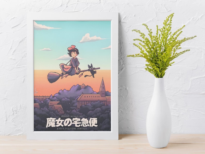 Op de afbeelding: Een ingelijste kunstdruk met een figuur met een rode strik die op een bezemsteel met een zwarte kat over een stad vliegt. De tekst luidt Hayao Miyazaki, Kiki's Delivery Service en de Japanse tekens 魔女の宅急便. Het is in een witte lijst.