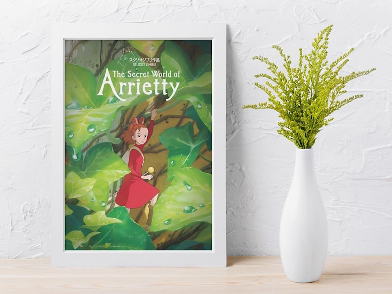 Pu&ograve; includere: Un poster incorniciato di "Arrietty - Il mondo segreto sotto il pavimento" con una ragazza in abito rosso tra grandi foglie verdi. Un vaso bianco contiene fogliame verde, su una parete bianca strutturata.