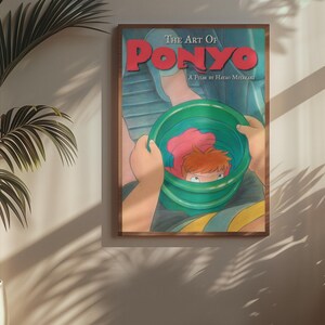 May include: A framed movie poster titled The Art Of Ponyo A Film By Hayao Miyazaki featuring a colorful illustration of a small character with orange hair inside a green bucket held by two hands against a blue background with red text.