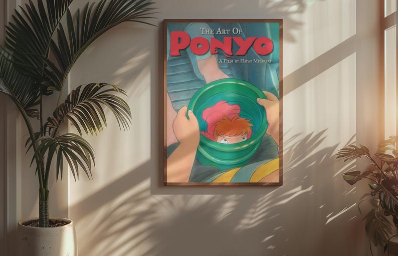 May include: A framed movie poster titled The Art Of Ponyo A Film By Hayao Miyazaki featuring a colorful illustration of a small character with orange hair inside a green bucket held by two hands against a blue background with red text.