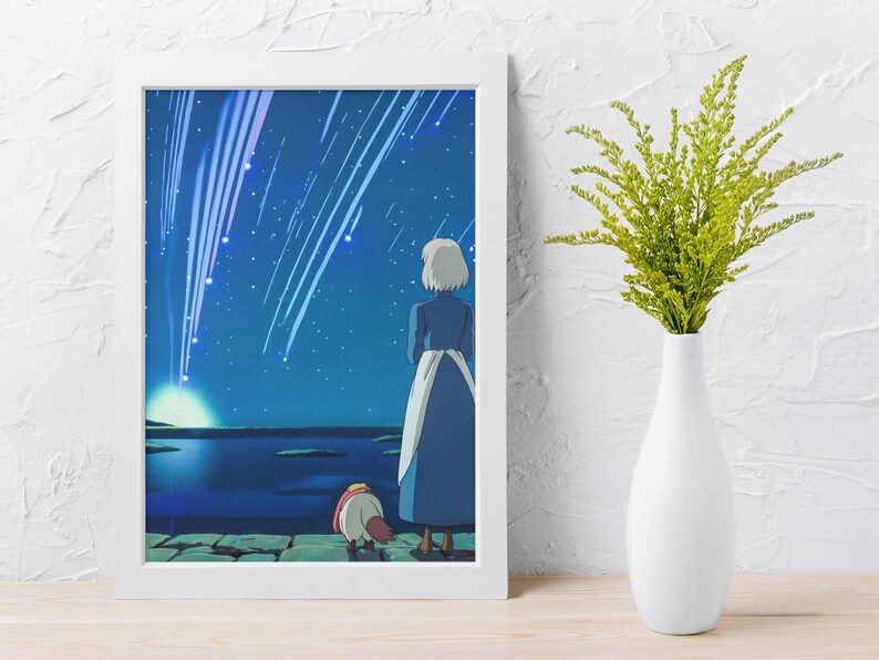 May include: A framed wall art print featuring an illustration of a person with short white hair in a blue dress and a small creature standing on a stone ledge, looking at a night sky filled with bright falling stars above a dark blue body of water.