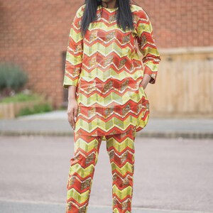 May include: A two-piece outfit featuring a long-sleeved top and matching pants. The ensemble showcases a vibrant chevron pattern in shades of yellow, orange, and red. The outfit is paired with white heels, creating a coordinated look.