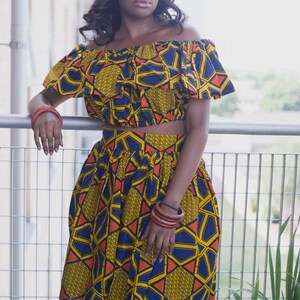 May include: A two-piece outfit featuring an off-the-shoulder crop top and a knee-length skirt. The ensemble showcases a vibrant geometric pattern in yellow, blue, and orange. The model is wearing red bracelets. The outfit is a bold statement piece.