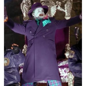 Può includere: Una figura che indossa un lungo cappotto doppiopetto viola, un cappello fedora viola abbinato e una cravatta verde. Il viso è dipinto di bianco con labbra rosse e capelli verdi. La figura si trova davanti a una sedia metallica decorata.
