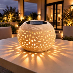 Op de afbeelding: Een bolvormige solar buitenlantaarn met een crèmekleurige buitenkant voorzien van een uitgesneden bladpatroon. Het bovenoppervlak bevat een zwart zonnepaneel. De lantaarn geeft een warm licht en werpt een sterpatroon op het oppervlak.