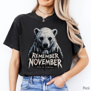 May include: A black short-sleeve t-shirt featuring a graphic of a white polar bear with dark eyes and a forest silhouette. The text reads REMEMBER NOVEMBER 2026 IS COMING in a stylized white font with red accents and glitch-like visual effects.
