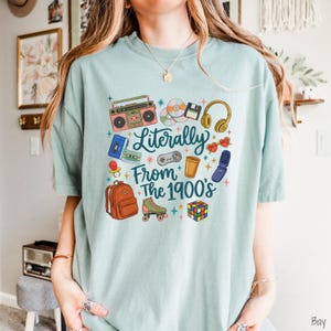 May include: A light green t-shirt featuring a graphic print with the text "Literally From The 1900's" surrounded by illustrations of a boombox, CDs, floppy disk, headphones, cassette tapes, game controller, cup, flip phone, backpack, roller skate, and cube.