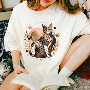 May include: An ivory t-shirt featuring a central graphic of a cat composed of various patterned segments including leopard print, polka dots, and geometric shapes. The cat is surrounded by a circular frame with gold spheres and pink leaves.