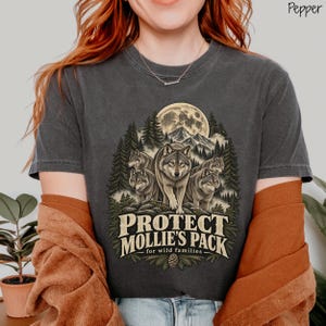 May include: A charcoal gray t-shirt featuring a graphic print of four wolves standing in a forest under a full moon. The text "PROTECT MOLLIE'S PACK for wild families" is printed in a cream-colored font below the wolves. The shirt has a crew neckline.
