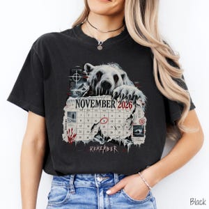 May include: A black t-shirt featuring a graphic of a white polar bear with red eyes peering over a torn calendar page for November 2026. The calendar highlights the 11th with a red circle and includes the word REMEMBER in a stylized font below.