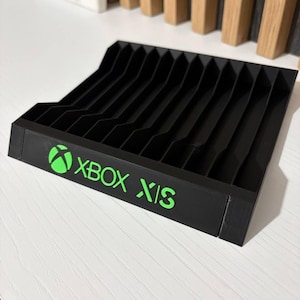 May include: Black Xbox X|S console stand with a series of vertical dividers. The stand has the Xbox logo and "XBOX X|S" in bright green lettering. The stand is made of a solid material and sits on a white surface.