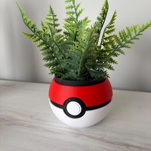 Könnte beinhalten: Ein dekorativer Pflanzentopf in Form eines roten, weißen und schwarzen Pokeballs, gefüllt mit grünen, farnartigen Kunstpflanzen. Der Pflanzentopf steht auf einer hellen Holzoberfläche.