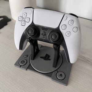 Può includere: Un controller di gioco wireless bianco e nero appoggiato su un supporto in plastica grigia. Il supporto presenta una base circolare con il logo PlayStation e due pannelli laterali quadrati con simboli di accensione ed espulsione.