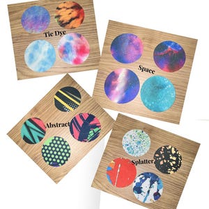 May include: Four wooden squares each display four circular designs. The designs include tie-dye, space, abstract, and splatter patterns. Each square has a different pattern theme, with the theme's name printed on the wood.