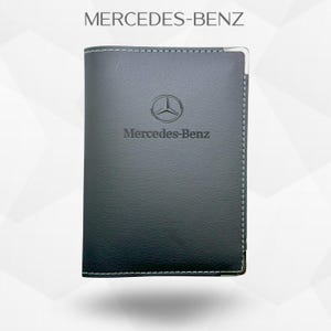 Puede incluir: Cartera de cuero gris oscuro con el logotipo y el texto Mercedes-Benz. La cartera tiene costuras blancas alrededor de los bordes y una esquina de metal plateado. El texto "MERCEDES-BENZ" está en la parte superior.