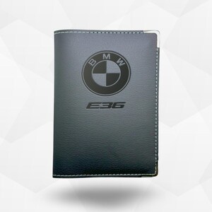 以下が含まれることがあります： 縁に白いステッチが入った濃いグレーの革製財布。BMWのロゴと「E36」の文字が前面にエンボス加工されています。財布にはシルバーのメタルコーナーが付いています。