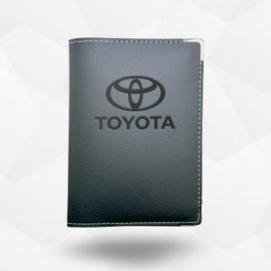 Pode incluir: Carteira de couro cinza escuro com o logotipo da Toyota em relevo na frente. A carteira tem costuras brancas nas bordas e um canto de metal prateado. A palavra "TOYOTA" está impressa abaixo.