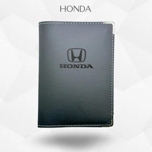 Puede incluir: Cartera de cuero gris oscuro con el logotipo de Honda en relieve en la parte delantera. La cartera tiene costuras blancas alrededor de los bordes y una esquina de metal plateado. La palabra "HONDA" está impresa en la parte superior.