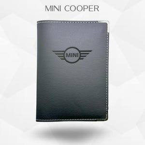 Peut inclure: Un portefeuille en cuir gris foncé avec le logo "MINI" en relief. Le portefeuille présente des coutures blanches et un coin en métal argenté. Le texte "MINI COOPER" est en haut.