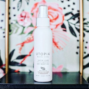 May include: White bottle of Utopia Bath Itd. Satin Luxe Moisturizing Dry Body Oil with a botanical toasted coconut and sandalwood scent. The bottle is 4 oz / 113.4 g.
