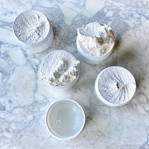 May include: Five white containers with a white, creamy substance inside. The containers are on a white marble surface.