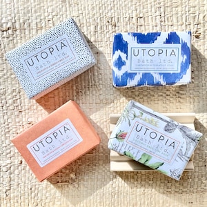 May include: Four bars of handmade soap with different scents and designs. The soaps are wrapped in paper with black and white polka dots, blue and white ikat, orange, and white floral patterns. The text on the soaps reads "Utopia Bath ltd." and the scent of each soap.