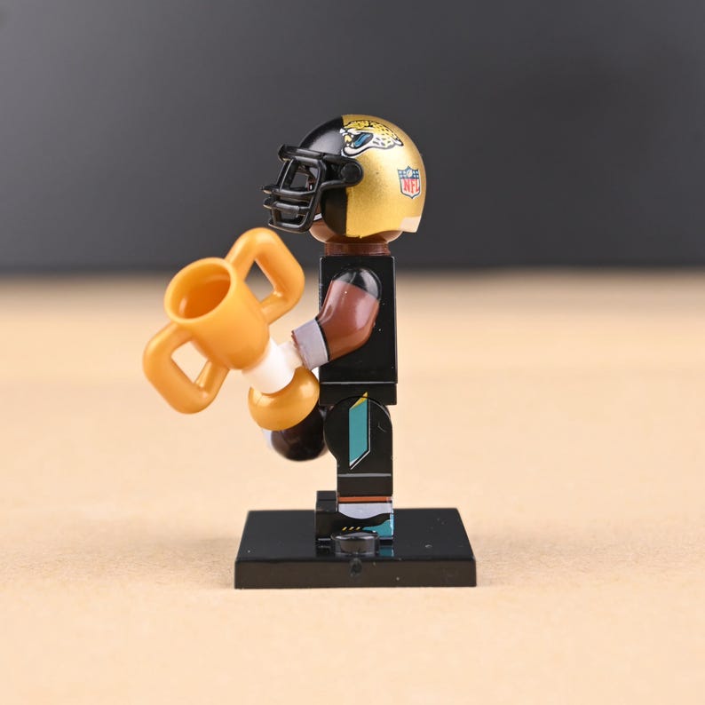 May include: A miniature football player figurine wearing a gold and black helmet with a black facemask. The figurine is holding a gold trophy. The player is wearing a black uniform with teal accents and is standing on a black base.
