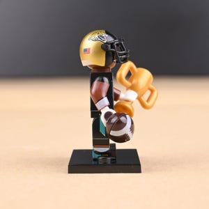 May include: A miniature football player figurine, wearing a gold and black helmet with a jaguar logo and an American flag. The player holds a football and a gold trophy. The figure is on a black base.