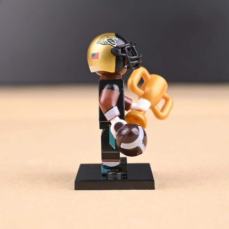 May include: A miniature football player figurine, wearing a gold and black helmet with a jaguar logo and an American flag. The player holds a football and a gold trophy. The figure is on a black base.