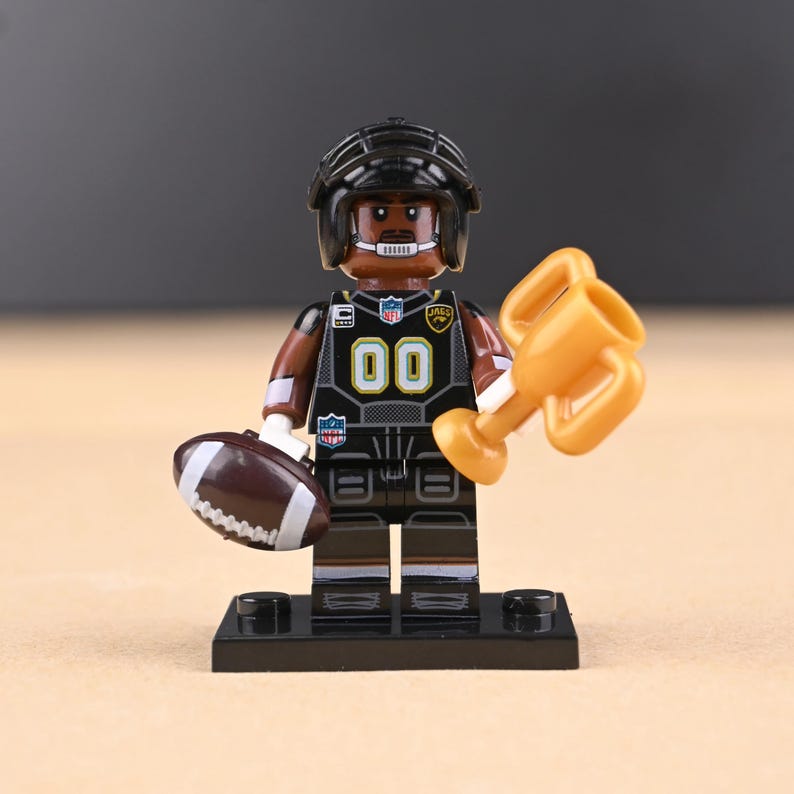 May include: A black and brown football player figurine with the number 00 on the jersey, holding a brown football and a gold trophy. The figurine is wearing a black helmet and is on a black base.