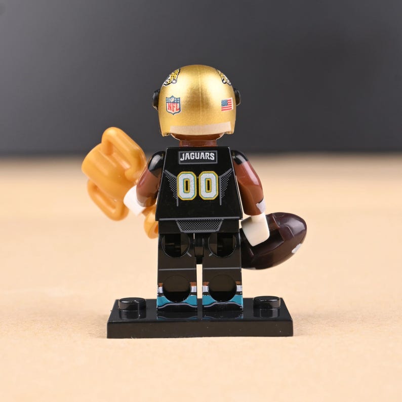May include: A miniature football player figurine, wearing a gold helmet and a black jersey with the word "Jaguars" and the number "00". The figure is holding a football and a trophy. The figurine is on a black base.