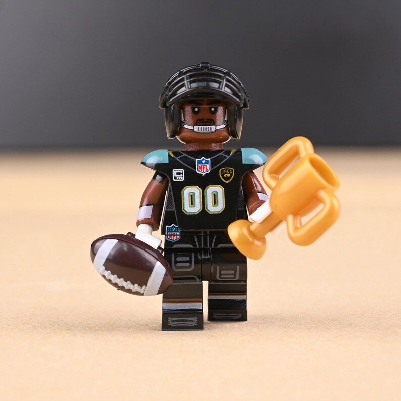 May include: A black and teal football player figurine wearing a helmet and uniform with the number 00. The figurine holds a brown football and a gold trophy. The uniform has the NFL logo.